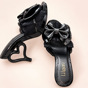 Black Lolita slip-on heels with glossy bow, black lace and simple hollow heart-shaped heel, on a light studio background.